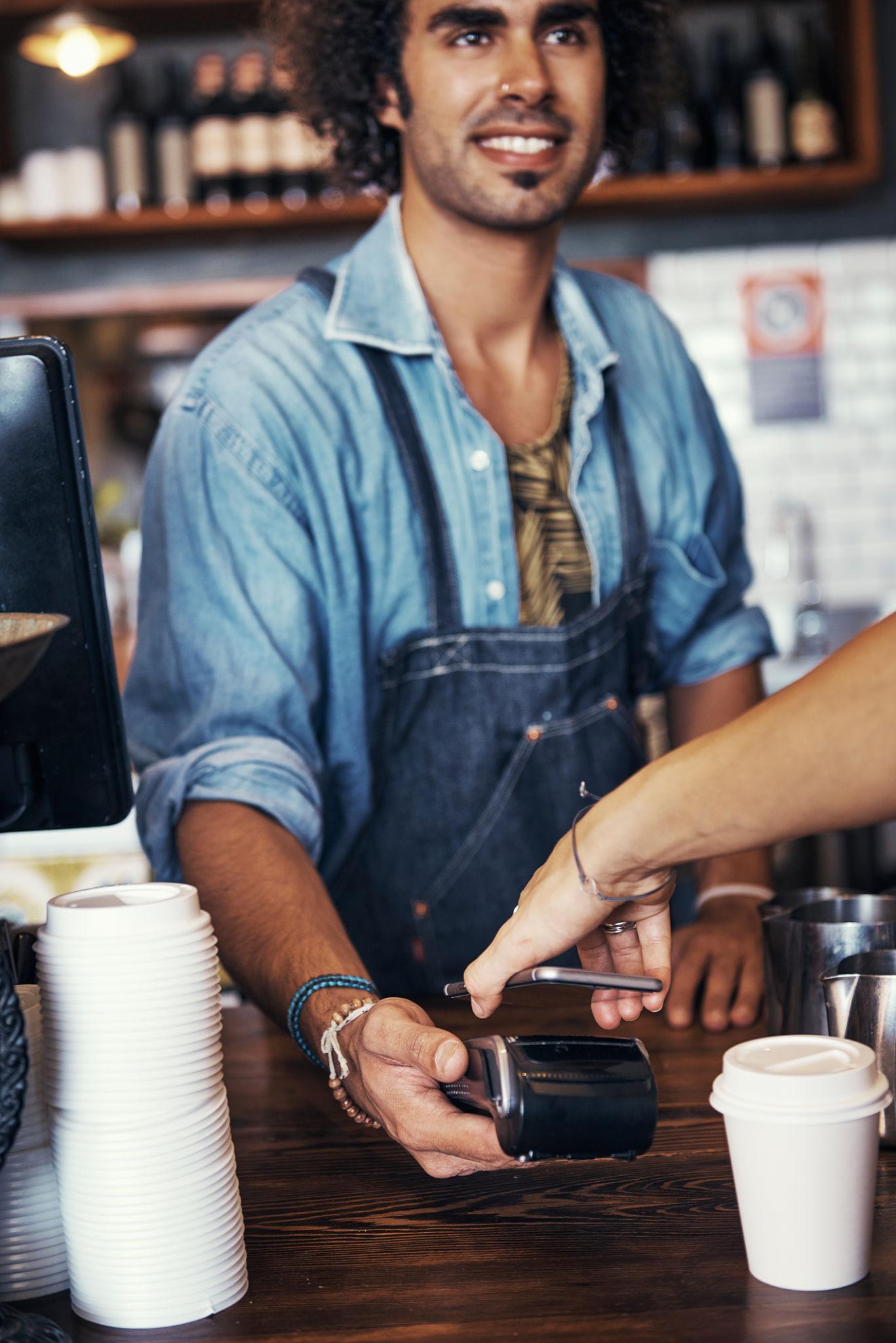 Cafe staff member at the counter handing a payment terminal to a customer.